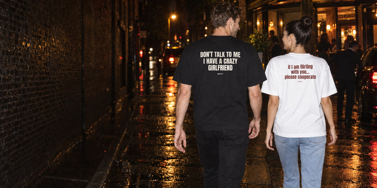 Theo and Mara walking away down a night street, each in an SBTXT tee with a sentence on the back